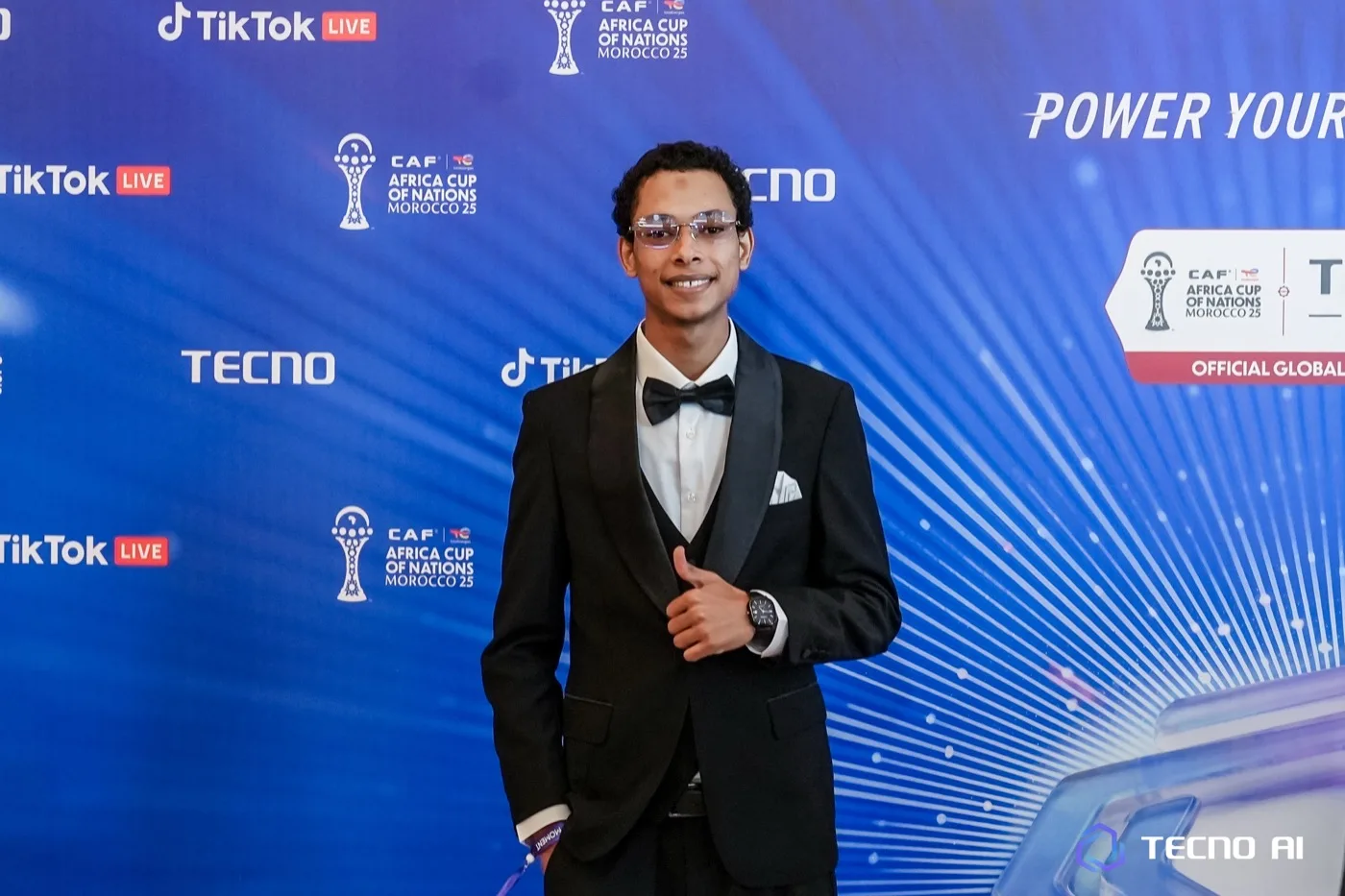 Le fondateur d'UniversH en smoking, pouce levé, sur le tapis rouge de la TECNO Glory Night, devant le mur officiel TECNO × TikTok Live de la CAN Maroc 25